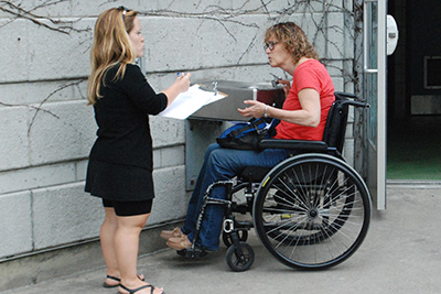 Lors d’une marche exploratoire, une femme en fauteuil roulant teste l’accessibilité d’un abreuvoir pendant qu’une femme de petite taille prend des notes.