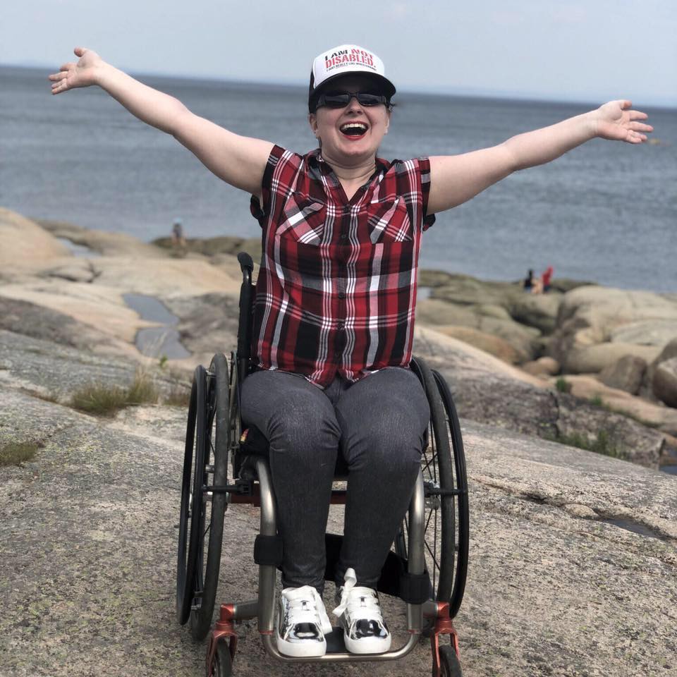 Description de l'image : Claudia Duchesne, en fauteuil roulant, fait un large sourire à la caméra, les bras écartés. Elle porte une casquette et se trouve sur des rochers, près d'une grande étendue d'eau..