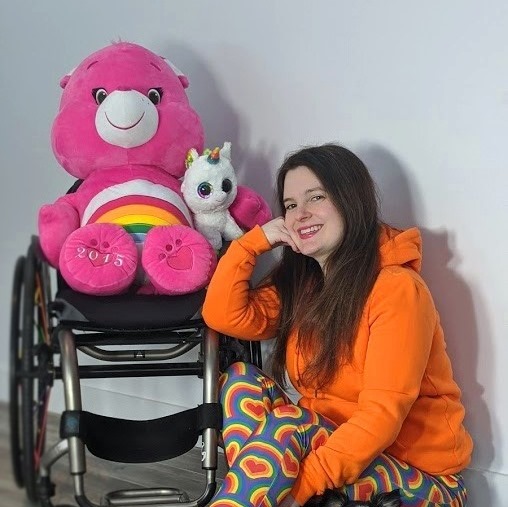 Description de l'image : Portant un coton ouaté orange et des leggings fluos, Claudia Duchesne sourit à la caméra, appuyée sur un mur. À ses côtés, on aperçoit son fauteuil roulant où des peluches colorés trônent.