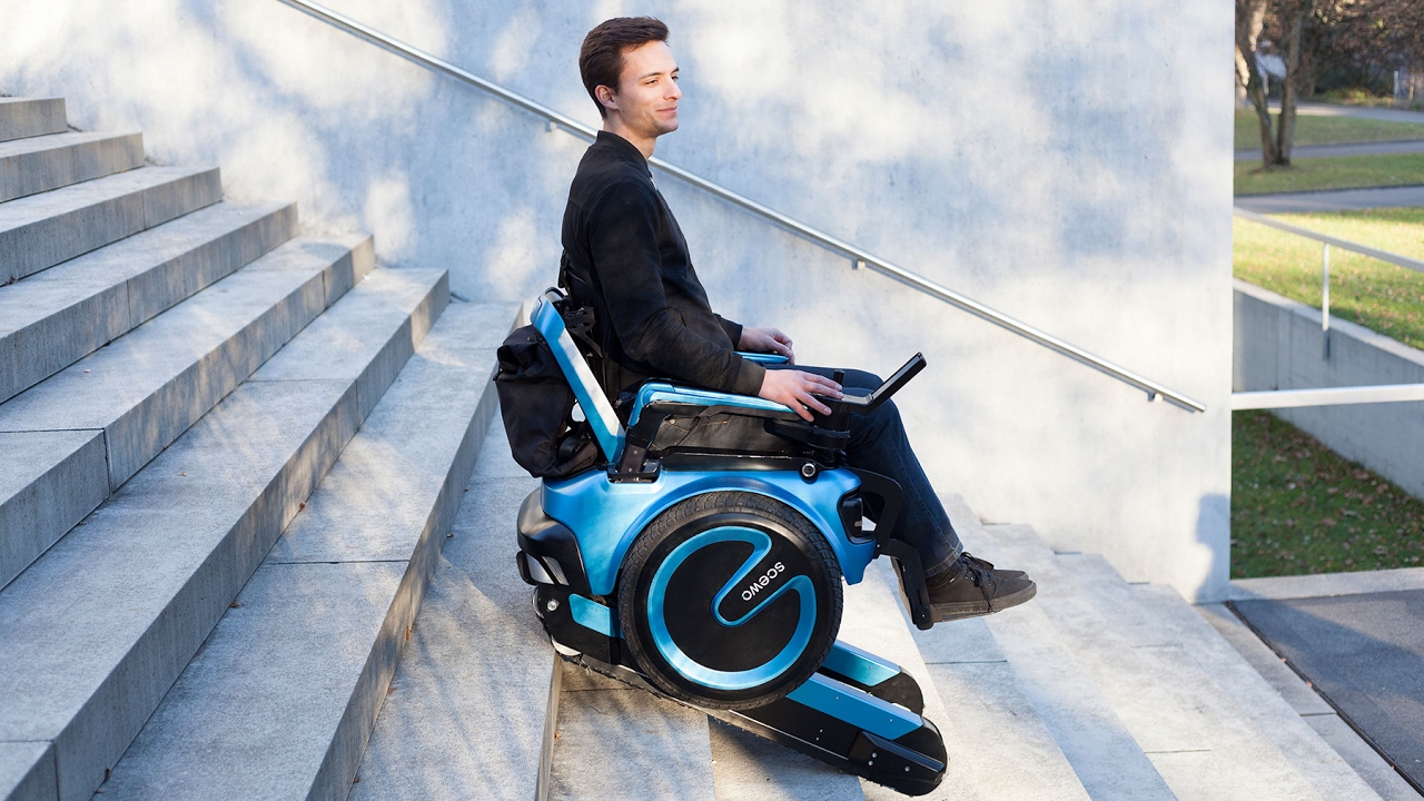 Description de l'image : Un homme monte les escaliers grâces aux chenilles mécaniques de son fauteuil électrique bleu.