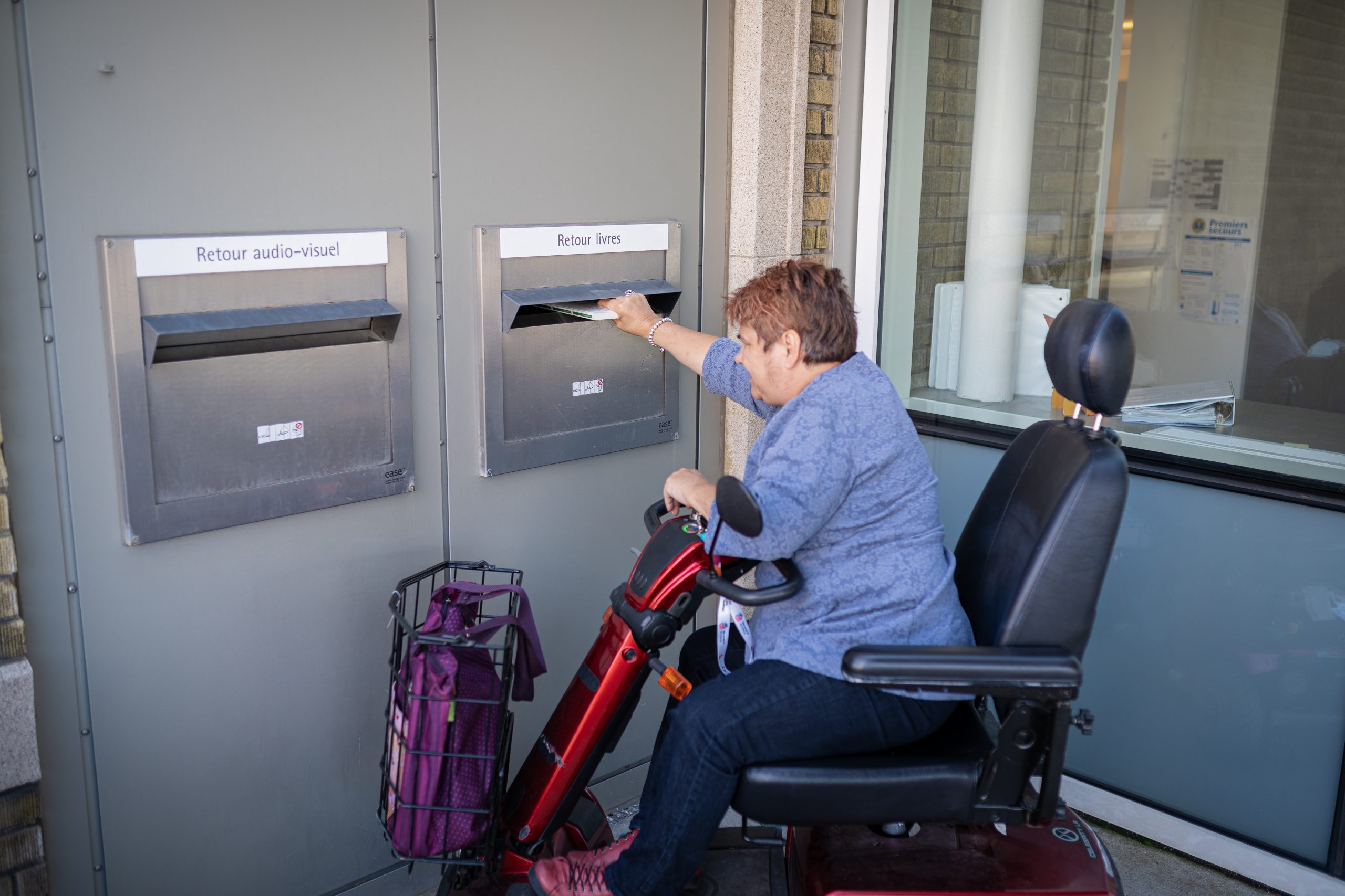 Une personne se déplaçant en fauteuil roulant électrique retourne un livre à une bibliothèque par une chute à livres extérieure.